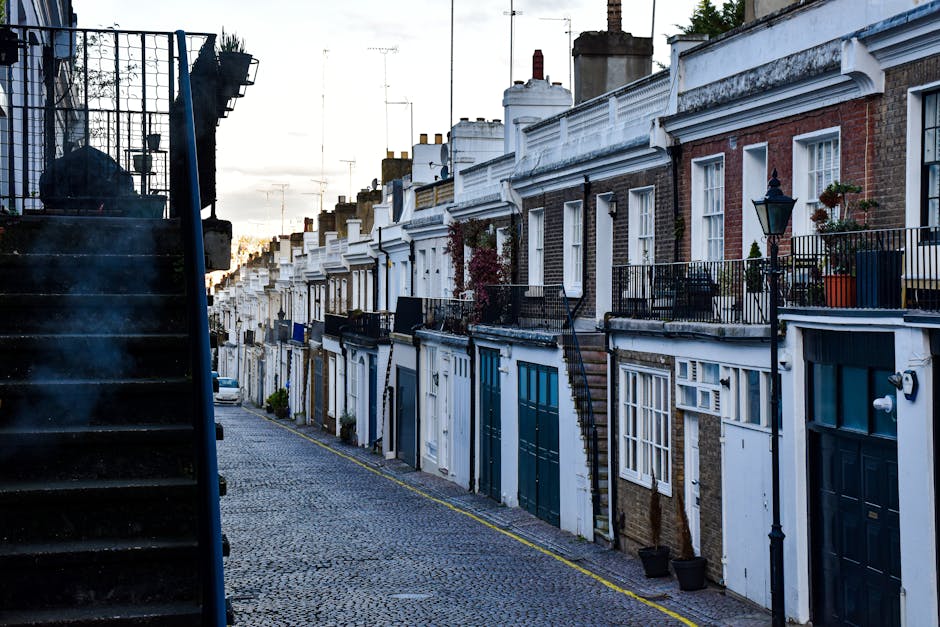 The image depicts a narrow residential street in Islington, with a row of terraced houses featuring classic brick facades, white window frames, and small balconies with black wrought iron railings, some adorned with potted plants. On the left side, a black metal staircase leads up to a building entrance, with dark steps and railing visible against the overcast sky that casts soft, diffused light on the scene. The cobbled pavement runs along the front of the houses, and a yellow line marks the edge of the parking area. A few parked cars are visible in the distance. The scene appears quiet, demonstrating a typical urban setting suitable for house removals, with no visible moving activity but clearly illustrating a location where furniture transport, packing, and home relocation services by Movers Islington could be carried out efficiently based on the environment.