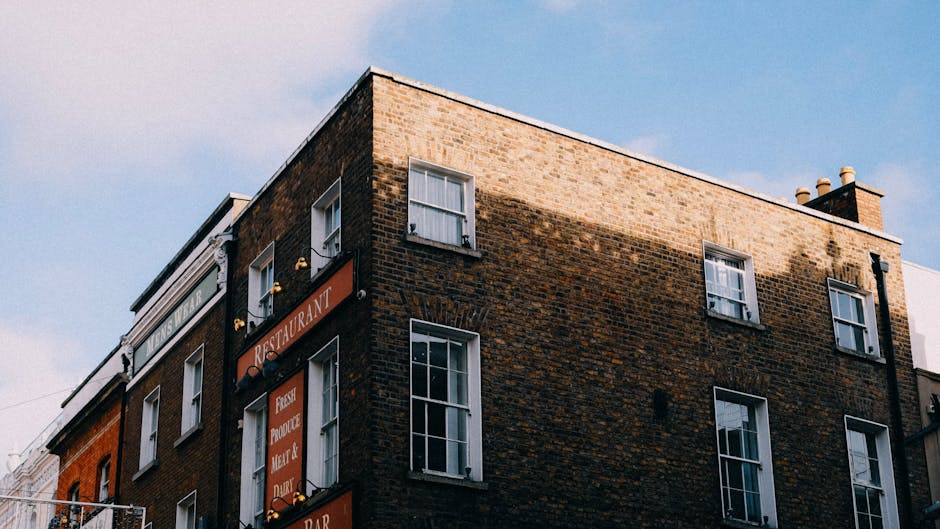 A multi-storey brick building located on a street corner in Islington, London, with visible windows and signage indicating a restaurant and bar. Above the establishment, the upper floors are plain brickwork with several sash windows, some open, revealing curtains. The roof has chimneys and ventilation pipes. The sky above is clear with a few wispy clouds, and the building's facade is partially lit by sunlight, creating shadows. In relation to house removals, a delivery area or loading zone may be inferred nearby, where furniture or boxes could be prepared for transport. Such environments are typical in urban settings where professional movers, such as Movers Islington, handle home relocation tasks, including furniture transport, packing, and logistics for flats situated above commercial premises.