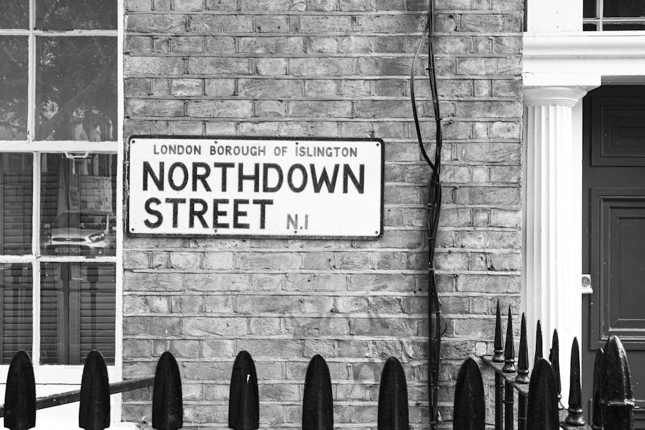 A black and white photograph of a residential exterior showing a brick wall with a street nameplate reading 'Northdown Street, Islington.' To the left, a window reveals a glimpse of interior furnishings and a parked car outside, suggesting the scene is in a city environment. Adjacent to the right, a decorative white column frames a dark door, with a set of black metal fence spikes in the foreground. Electrical wires are visible, running vertically along the brick wall. The image is taken during daylight, with natural light illuminating the scene evenly. This photo, associated with house removals and relocation services provided by Movers Islington, captures the typical urban setting where furniture, boxes, and packing materials might be staged for loading onto a van, possibly during a home relocation or moving process. It emphasizes the outdoor environment of a property and the logistical considerations involved in furniture transport within an urban area. The scene is free from human activity, focusing on the structural and environmental context of moving logistics, ideal for illustrating urban house removal services or moving processes in Islington.
