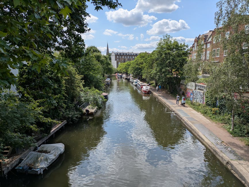 A canal scene in a residential area of Islington, with calm water reflecting the partly cloudy sky above. On the left, lush green trees overhang the water, and a small boat covered with a protective tarp is moored close to the bank. On the right, a narrow footpath runs alongside the canal, with a few pedestrians walking and a line of parked cars visible further down the street. Multi-storey brick and stone buildings, typical of London architecture, line the right side of the canal, some with graffiti on their lower walls. Further in the background, a church spire rises above the treetops, indicating a historic local landmark. The scene captures a quiet moment in a picturesque urban setting, where [COMPANY_NAME] often assists with home relocation, furniture transport, and packing and moving services in the Islington area, with the canal serving as a scenic backdrop during residential moving projects.