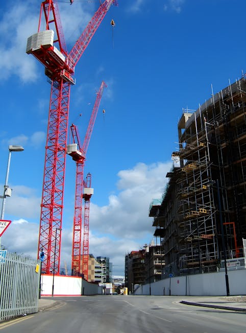 A construction site during daylight shows two large red tower cranes with long arms extending into a partly cloudy blue sky. The cranes are equipped with white counterweights and cables hanging from their jibs, used for lifting materials. To the right, there is an unfinished multi-storey building clad in scaffolding and safety netting, indicating ongoing construction work. The foreground features a paved road with a streetlight on the left and a metal fence along the perimeter of the site. In the background, other modern buildings and additional cranes can be seen, suggesting an urban development area. This image captures the heavy machinery and infrastructure involved in building projects, relevant to logistics and transportation for house removals and moving services, as performed by Movers Islington.