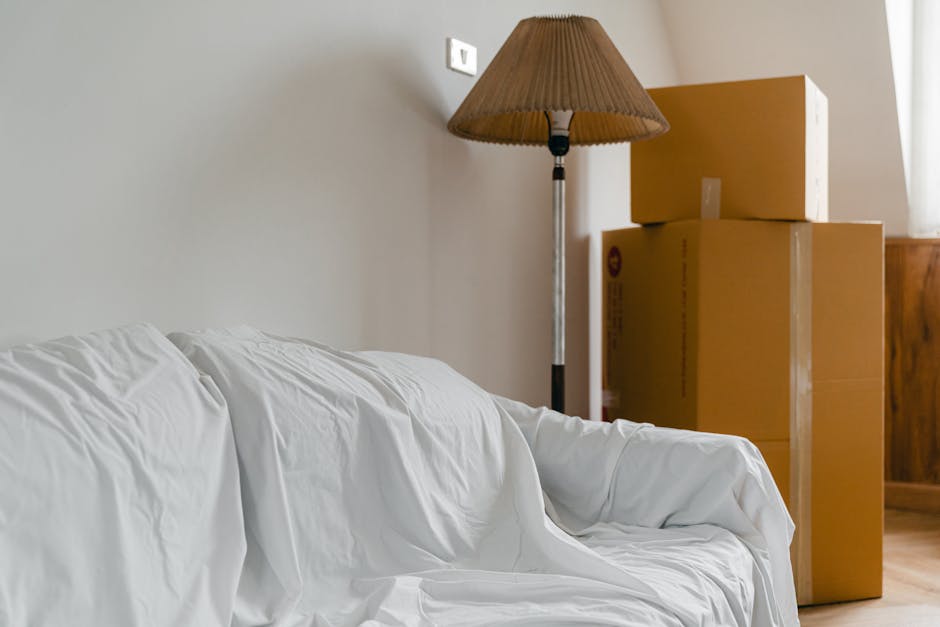 Inside a home, there are three cardboard moving boxes stacked beside a wooden cabinet near a window with natural light. A floor-standing lamp with a brown pleated lampshade is positioned next to the boxes. In the foreground, a white sofa with a loose cover is visible. The scene depicts part of a furniture packing and loading process, with boxes prepared for relocation. The environment appears tidy and well-lit, emphasizing the interior space prior to moving. This setting relates to house removals and furniture transport services as part of home relocation, with visible packing materials and furniture arranged for transportation. The image aligns with the content on Movers Islington's page about preparations before booking removals services.