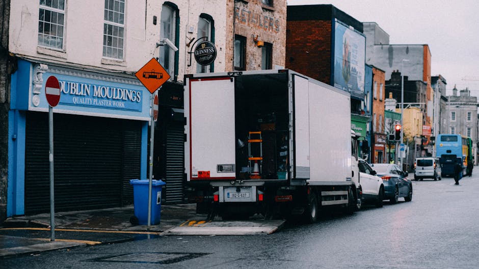 A street scene during a home relocation shows a narrow urban road flanked by multi-storey buildings with stone facades and black fire escapes. In the background, a white moving van branded with 'uniserve' and a black truck marked with 'Becton Transport' are parked, partially blocking the road, with a motorcycle also visible next to the vans. On the street, a man in a dark jacket and jeans is walking away from the camera, carrying a black bag over his shoulder. An older man wearing a dark coat and light pants is walking towards the camera, crossing the road. The scene is illuminated by natural daylight, highlighting the logistical activity involved in furniture transport and packing for house removals, as managed by professional services like Movers Islington.