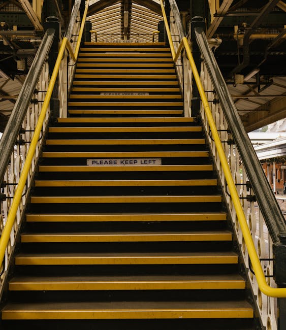 A set of broad industrial metal stairs with yellow safety handrails on both sides leads up to an elevated platform inside a building, possibly a warehouse or storage facility used for home relocation or furniture transport. The stairs are fitted with anti-slip yellow and black striped treads, and signage on the steps reads 'PLEASE KEEP LEFT', indicating a regulated moving or loading process. The environment is illuminated by overhead industrial lighting, revealing parts of supporting structures, steel beams, and a partially visible loading area at the top. This scene is part of a professional removals operation, where large items or boxes might be transported via the stairs for loading onto a vehicle, consistent with the services offered by Movers Islington for house removals and moving logistics.