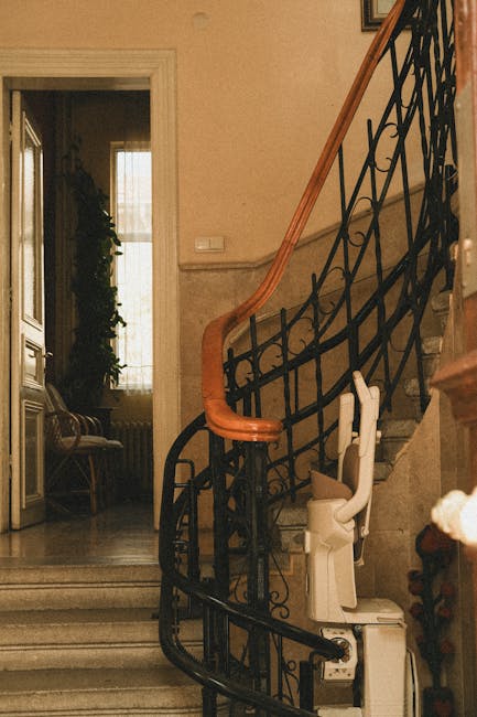 A spiral staircase with a black wrought iron railing and wooden handrail inside a residential property. Adjacent to the staircase is a white stairlift chair positioned on the stairs, indicating accessibility adaptations for moving within the home. To the left, a doorway with a doorframe leads to a room illuminated by natural light through a large window, partially draped with curtains. Inside the room, there is a wooden bench and a tall plant near the window. The staircase and surrounding environment feature a beige wall finish, with visible marble or stone steps. This setting exemplifies the internal environment of a house where furniture and equipment are being prepared for removal or relocation, consistent with professional services such as those offered by Movers Islington specializing in house and large item removals, especially where stair and lift access issues may need to be managed during the packing and moving process.