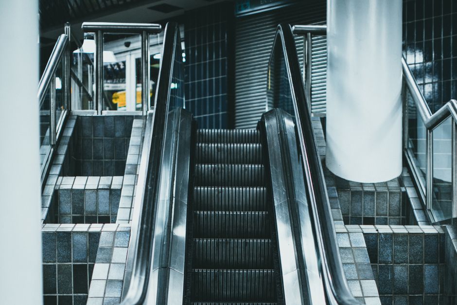 A set of broad industrial metal stairs with yellow safety handrails on both sides leads up to an elevated platform inside a building, possibly a warehouse or storage facility used for home relocation or furniture transport. The stairs are fitted with anti-slip yellow and black striped treads, and signage on the steps reads 'PLEASE KEEP LEFT', indicating a regulated moving or loading process. The environment is illuminated by overhead industrial lighting, revealing parts of supporting structures, steel beams, and a partially visible loading area at the top. This scene is part of a professional removals operation, where large items or boxes might be transported via the stairs for loading onto a vehicle, consistent with the services offered by Movers Islington for house removals and moving logistics.