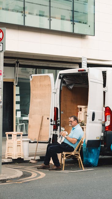 A man seated on a wooden chair outside a white delivery van during a home relocation process, with the van's rear doors open revealing interior space filled with cardboard boxes, wooden furniture pane