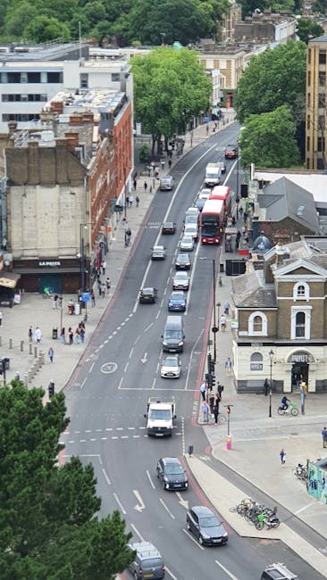 An aerial view of a city street exhibiting a line of parked cars along the curb, with moving vehicles including a red double-decker bus and several smaller cars traveling along the road. The street is bordered by mixed-use buildings, including a brick structure and a white building with a gabled roof. Sidewalks on both sides of the road are populated with pedestrians, some walking and others waiting at crosswalks. Green trees line the sidewalks, providing shade and greenery within the urban environment. In the foreground, a section of a blue bicycle parked next to a lamppost is visible, while further down, a small bicycle rack with parked bikes enhances the scene. The image captures the typical busy atmosphere of a central London neighbourhood, illustrating aspects of urban transport and street life, relevant to house removals, furniture transport, and moving logistics, as related to the services offered by Movers Islington.