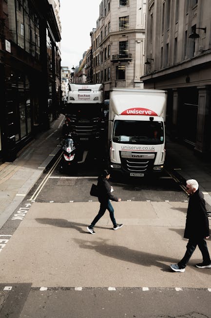 A narrow urban street scene showing two white delivery vans parked on the road, with one branded 'Uniserve' and the other 'Becton Transport.' The vans are positioned side by side, slightly offset, with the 'Uniserve' van closer to the camera. In front of the vans, a black scooter is parked alongside the curb, facing away from the vans. Pedestrians are crossing the street: a woman wearing a black jacket and carrying a backpack is walking from left to right, while an older man with white hair, dressed in a dark coat, is walking in the opposite direction. The street is flanked by tall, light-colored buildings with multiple windows and fire escapes, with the scene showing a typical city environment. The photo was taken during daylight hours, capturing the loading process involved in home relocation or furniture transport, possibly linked to a house removals service such as Movers Islington, operating in the area around Upper Street in Islington.