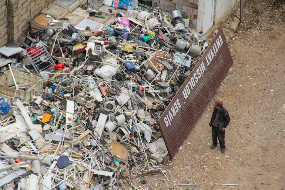 A large pile of discarded household items and waste materials, including broken appliances, metal frames, plastic containers, cardboard boxes, and various debris, located outdoors next to a closed bro